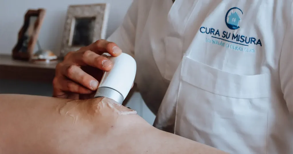 Ultrasuonoterapia a domicilio in Lombardia: un professionista della riabilitazione esegue un trattamento con manipolo a ultrasuoni e gel conduttore sul ginocchio del paziente direttamente a casa.